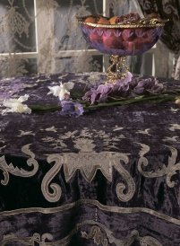 The picture shows a light-colored wooden bench with two yellow-and-white throw pillows on it. The bench is placed in front of an arch-shaped window, and there is a large mirror on the wall behind it. There appears to be some kind of greenery outside the window, as well as a star patterned curtain. The room is decorated with various decorative items, including a framed drawing, a vase of flowers, and a shelf of books outlined in gold.