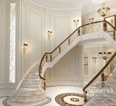 This picture shows a luxurious, ornate staircase with a white and gold railing. The stairs are made of rich, dark wood, and each step is highlighted with a contrasting white edging. The top of the stairs is lined with a grand black and white marble floor, surrounded by magnificent pillars topped with marble capitals. On either side of the staircase is an ornate white and gold gilded wall featuring an intricately designed geometric pattern.