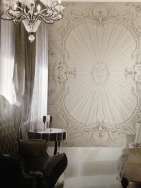 This picture shows a luxurious, elegant dining room with an intricately carved white ceiling, marble flooring, and tall arched windows. The walls have deep brown paneling and are adorned with various pieces of art. A large, ornate chandelier hangs from the center of the room over the round dining table, which is surrounded by eight white chairs with plush velvet upholstery. An assortment of vibrant flowers decorates the table in a crystal vase, providing a lush centerpiece.