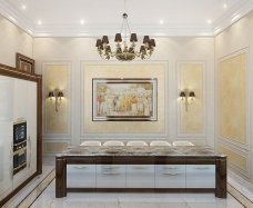 This is a picture of a luxurious dining room designed by Antonovich Design. The room features an ornately carved light wooden table, surrounded by white upholstered chairs and colorful throw pillows. A large crystal chandelier hangs from the ceiling, and a luxurious area rug covers the floor. The walls are painted in a light yellow hue and decorated with large gold framed mirrors.