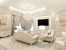 The picture depicts a luxurious living room with an ornate gold-and-white ceiling and large, luxurious chandelier. The walls are painted in a light gray hue and adorned with intricate white crown molding. The room features high vaulted ceilings, ample natural lighting from several tall windows, and a large white marble fireplace. The floor is covered in a light tan carpet and the furniture consists of two beige sofas, a large armchair, and a matching ottoman. Other accessories include a round coffee table and a pair of decorative vases.