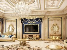 This picture is of an elegant interior living room design. It includes a white U-shaped sectional sofa, a matching accent chair, and a round ottoman with intricate patterned upholstery in gold and cream tones. On the walls, there are two large abstract paintings in creamy gold and white, while the ceiling is adorned with a brass and crystal chandelier. The floor is composed of two different light wood grains, creating a subtle yet stylish contrast. The overall effect is an inviting, luxury interior space.