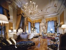 This picture shows a luxurious and modern living room interior with a large, cream-colored couch set against a neutral gray wall. The walls feature an arched design detail in a dark wood finish, while the floor is covered with a light-colored rug. Across from the couch is an elaborate gold and white coffee table with matching armchairs. A few small plants accent the furniture pieces, and two differently-sized chandeliers hang from the ceiling to provide a warm ambience.