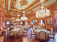 The picture shows an elegant and modern dining room. The walls are beige with golden accents, the floor is marble, and a crystal chandelier hangs from the ceiling. There is an eye-catching dining table in the center of the room with six upholstered chairs. The table has a white tabletop and gold legs. A buffet also stands in the corner of the room, with art and decorative pieces adorning the surface.