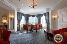 This picture shows a luxurious, white and gold sofa surrounded by a warmly lit room. The furniture in the room has a map motif, with gold and beige armchairs, an intricately designed side table, and a decorative mirror above the couch. The walls are a light shade of beige, with a few white accents, and the floor is wood-grained. There is a large rug, patterned with various symbols, covering the majority of the floor. The fireplace, across from the sofa, is made of light stone and has a wooden mantle.