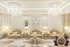 This picture shows a modern and luxurious living room. The room features two white sofas with gold detailing, a round glass coffee table, and an intricately designed white grand piano. The walls are decorated with abstract art pieces and the room is lit by a large crystal chandelier and wall sconces. The flooring is dark hardwood and there is a tray ceiling with recessed lighting.