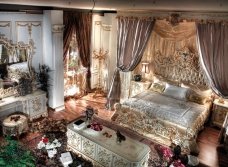 This picture shows a luxurious bedroom in an opulent home setting. The bedroom has a large canopy bed with a rich gold and velvet-textured headboard, deep crimson and gold bedding, and a deep tufted ottoman at the foot of the bed. A richly patterned rug sits underneath them both, while a set of floor-to-ceiling dark gold drapes provide privacy along the windows. The walls are painted a soft, pale yellow, and the room is finished off with a crystal chandelier and some art deco lighting fixtures.