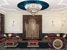 This picture shows a luxurious living room with a curved shaped wall of light beige marble and a circular floor inlay. On the marble wall is an ornate gold-framed mirror and two black-and-white portrait paintings. At the center of the floor inlay is a circular table and four plush armchairs upholstered in white fabric, surrounded by a colorful Persian rug. There are two modern floor lamps and two tall, classical white column lamps that flank the center table. The room is completed with two boutique sofas and two small round tables, along with a