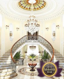 This picture shows a grand and luxurious foyer with high ceilings and decorated with stunning crystal chandeliers. The walls are painted in a neutral beige tone, with accents of gold detailing on the trim, door frames, and door handles. The floor is a dark wood look with an intricate tile medallion in the centre and a patterned marble floor leading to the grand staircase. To the side you can see a sitting area with white furniture and a large gold framed wall mirror.