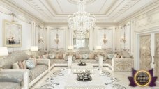 This picture shows an ornate and luxurious bathroom. It features a large, white ornate sink on a marble countertop, a gold-accented mirror above the sink, two white curved cabinets with gold handles, a large golden wall sconce, a white stone wall with a gold trim, and a white marble floor.