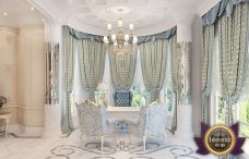 This picture shows an interior design of a luxurious bedroom. The room features a white and gold color palette, with gold accents throughout, including the canopy-style bed frame, crystal chandelier, and multiple armchairs and a table in the corner of the room. A large oval shaped mirror hangs above the bed, and there is also a white rug on the floor. The walls are lined with white, built-in shelves, adding a touch of elegance to the space.