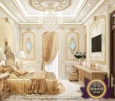 This is a luxurious bedroom design by Antonovich Design, located in Dubai, UAE. It features tall and elegant tufted upholstered walls, a richly decorated ceiling with dark wood trims and an impressive crystal chandelier, an ornate headboard with gold accents, plush bedding, and elegant furniture pieces such as nightstands, a chest of drawers, and an upholstered chaise lounge. The overall look is one of luxury and sophistication.