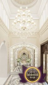 This picture depicts a luxurious white and gold bedroom. The room features a large canopy bed with intricate carved details and an ornate headboard. The bed is draped in luxurious white bedding and several gold pillows. On either side of the bed is a white nightstand with golden accents. Along the far wall of the room is a beautiful white armchair with a matching ottoman and a tall gold lamp. Above the bed is a white and gold chandelier and two decorative wall sconces.