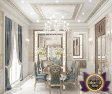 This picture shows a classic and luxurious living room featuring an ornate two-tone coffee table, beige sofa, and velvet chairs. The walls are painted in a rich cream color with decorative moldings, and the window is framed in a classic gold trim. A pair of black lamps add to the sophistication of the room and create a warm ambiance. A classic Persian rug ties the design together.