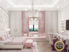 The picture shows a luxurious bedroom designed in a modern style. The bedroom features a large four-poster bed with gold accents and matching golden curtains draped over the bed frame. The walls are covered in white silk fabric with a gold pattern, adding to the regal atmosphere of the room. The floors are a coordinating light wood and there is a cozy seating area featuring a beige velvet sofa and chair. In the center of the room is a round table with an intricately detailed base and topped with a framed abstract painting.