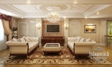 This picture shows an ornate and luxurious living room. The walls are a deep eggplant purple and the floors are covered in an intricate light beige carpet. The room features a white and grey sofa set, a glass coffee table adorned with gold accents, and two tall armchairs with patterned cushioning. A sparkling chandelier hangs from the ceiling and two large windows provide a view of the outside.