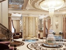 This picture shows an ornately decorated hallway with a luxurious crystal chandelier that is hanging in the center. The walls are painted a deep shade of red, and have intricate gold leaf detailing. There is a marble floor, and there are several decorative accents around the room, such as vases and statues. The ceiling is high and has a beautiful coffered design in the center.