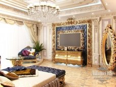 This picture shows an elegant and luxurious living room in a home. The room features a large, patterned blue and white sofa with gold accents, two cream-colored armchairs with decorative throw pillows, a white glass coffee table with a large golden centerpiece, and a blue and white rug on the wooden floor. The walls are painted light blue and decorated with art pieces and white curtains. The lighting is provided by a modern crystal chandelier overhead.