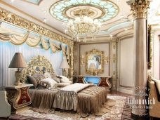 This is a picture of a modern bedroom interior design. It features a large four-poster bed with white sheer curtains and a light green bedspread. The walls have a greyish-green color and the floor is made up of a light brown hardwood. There are two nightstands on either side of the bed, each with lamps, and one smaller table in the corner with a vase and flowers. On the wall next to the bed is a large art piece with different shades of blue and hints of gold.