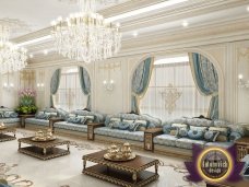 This picture shows a luxurious dining room, whose walls, ceiling and floor are all done in a white textured marble. In the center of the room is a modern rectangular wooden dining table with two upholstered chairs. The space also includes a dark wood credenza against the wall for added storage, with a modern vase of white flowers on top. On the opposite wall is a large gilded mirror and a framed work of art. Small sconces line the walls, providing ambient lighting to the room.