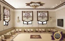 This picture shows a luxurious living room with a large central sofa in a sandy beige color. The walls are painted a light grey, and the floor is covered with a plush white rug. On the walls there are beige curtains, and several ornate gold framed paintings. The ceiling is adorned with a traditional chandelier, and on either end of the sofa are two cream-colored armchairs with small circular side tables between them. Finally, a light wooden coffee table and two matching end tables are present in the center of the room.