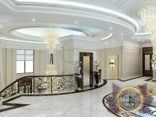 This picture shows a curved wooden staircase placed in the center of a room with ornately decorated white walls. The staircase has intricate carvings on the railings and steps, and is made from high-quality wood. On the walls are large wall fixtures, with two standing lamps on either side of the stairs. The room has a rich and luxurious aesthetic, with elegant décor and fine craftsmanship.