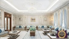 This picture shows a luxurious dining room with stone floors, gold accent walls, and a white ceiling. An ornate chandelier hangs from the ceiling, and a large wooden table is surrounded by upholstered chairs. A lighted buffet cabinet is placed against the far wall, and a large decorative mirror hangs above it. There is an elegant rug beneath the table, and a large painting hangs on the wall above the buffet.