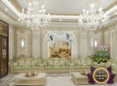 This picture shows a beautifully designed white and gold colored interior in a sitting area, featuring a comfortable sofa with accent pillows and two velvet armchairs, all placed on a light gray rug. The focal point of the room is a circular gold-framed wall mirror and a golden side table with a glossy faux marble top. An oversized abstract painting hangs above the sofa, and there are several tall plants scattered around the space to add color and texture. Overall, the space has a luxurious and modern feel.