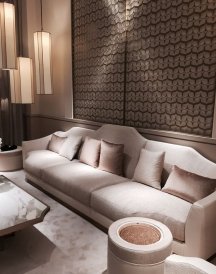 This picture shows a luxurious and modern living room with an exquisite design. The walls are a light cream color, with a soft texture. The ceiling is a more daring hue of blue, with some gold accents around the edges. In the centre of the room, there is a white leather sectional sofa surrounded by two round coffee tables and two armchairs, all atop a beautiful patterned rug. The room is also adorned with gold and crystal chandeliers, as well as several pieces of artwork hung on the walls.