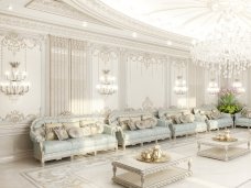 This picture shows a modern luxury living room with a white and gold color palette. The room features a white carpet, white furniture, and a gold chandelier. There is also a wall of built-in shelves in a light wood finish and a large floor-to-ceiling window that brings in plenty of natural light. The room also has several art pieces including a large abstract painting and a sculpture on the coffee table.