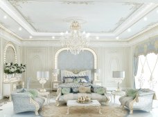 This picture shows a luxurious and modern bedroom. The room is decorated in a contemporary style, with an accent wall featuring a light brown wood-like texture. The room is furnished with a large, upholstered light grey headboard, two nightstands, and a matching chest of drawers. The bed is dressed in white linens and pillows, and a plush area rug covers the floor. Light fixtures on either side of the bed provide additional light to the room.