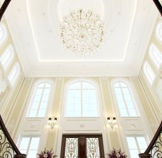 The picture is of a luxury living room in a mansion. It features a light gray sectional sofa with upholstered arm rests and white pillows, an ornate fireplace with a large white mantel, an ivory tufted ottoman, two light gray chairs with tufted backs, a round wooden coffee table, an ornate crystal chandelier, and neutral colored walls with abstract artwork.