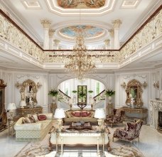 This picture shows an elegant dining space in a contemporary style. There is a marble dining table in the center of the room that is surrounded by cream-colored tufted chairs with golden metal legs. The walls are adorned with a luxurious mural and detailed accents, and there is a glass chandelier that hangs from the ceiling. The floor is tiled in a pattern of soft beige and brown.