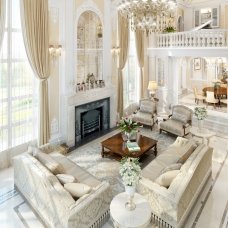 The picture shows a marble clad luxury bathroom with a large glass enclosed bathtub nestled at the center of the room. On either side of the tub are two large built-in shelves that display a collection of white towels and spa products. The walls and floor of the room feature white marble tiling, with two wood vanity units on each side of the space. Above the tub is a sparkly ceiling light fixture and recessed lighting accents the warm marble atmosphere.