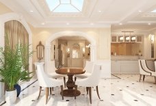 This picture shows a luxurious modern kitchen with a large island in the center. The counters are made of white marble, while the floors are white porcelain tile. There are curved wooden cabinets around the perimeter of the room, as well as a dining table and chairs. The walls and ceiling are decorated with intricate wall and ceiling moldings, and there is a large vent hood overhead. A large window looks out onto a lush garden.