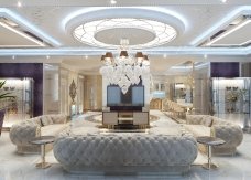 This picture shows a luxurious and modern living room with high vaulted ceilings. The room features cream colored walls, floor-to-ceiling windows, and a marble fireplace. The room also has a distinctively modern black leather sofa, two matching armchairs, and a white coffee table with glass shelves. There are also two art prints hanging on the walls, an ornate chandelier hanging from the ceiling, and several potted plants around the room.