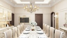 This picture shows an ornately decorated dining room with a large rectangular table surrounded by 10 chairs. The table has a marble top with a carved pedestal base and is adorned with elaborate centerpieces and candelabras. The walls are covered in a soft yellow damask wallpaper, which is complemented by white wainscoting and a dark wooden floor. The neutral tones are accented by the bright blue velvet curtains that frame the windows.