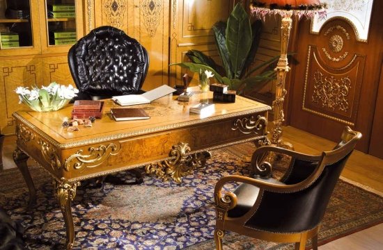 This picture shows a modern living room with a navy blue and beige color palette. The walls are painted in a deep navy blue, while the sofa and other accent furniture are upholstered in beige. The floor is covered in a light gray rug, and there is also a tufted ottoman in the center of the room. The decor features geometric patterns, including pillows on the sofa and a wall-mounted art piece with white lines.