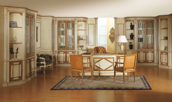 This picture is of a luxurious living room with high vaulted ceilings and a balcony overlooking an outdoor pool area. The walls are covered in a warm beige wallpaper and feature elegant crown molding. A large crystal chandelier hangs from the center of the room and provides a soft, ambient light. The furniture is cream-colored and includes two armchairs, a sofa, and a long coffee table with a glass top. On the coffee table, there is a vase of white flowers and books.