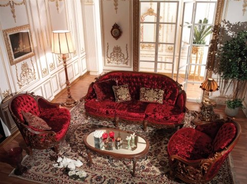This picture shows an ornate and luxurious living room with high ceilings and wall-to-wall windows. The dark maroon walls are complemented by velvet-like furniture, gilded gold accents, and a round chandelier. A white marble fireplace stands against one of the walls, while a patterned area rug lies on the floor. The room is framed by two beige armchairs and a glass coffee table, complete with white flowers and art books.