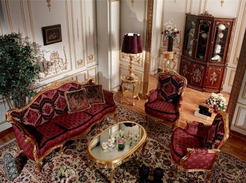 This picture is showing a luxurious modern-style bedroom in an opulent home. The bed has a grand frame upholstered with a bright, silky-looking material and flanked on either side by two tall glass lamps. The bed is layered with large white pillows, and a gray blanket that adds texture to the scene. The walls are painted a light gray with white trim and adorned with a golden framed landscape painting which creates a contrast against the neutral colors. The room also features a round wooden side table with a stylish black lamp, a large cream-colored armchair with detailed