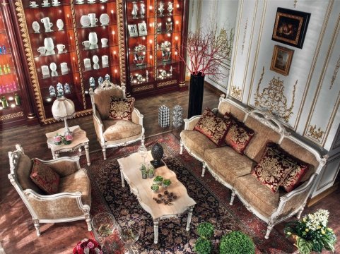 This picture is of a luxury living room. The room is decorated with luxurious designs, including a rich red velvet couch, a gold and glass coffee table, and brown, cushioned armchairs. There is an ornate chandelier hanging above the room, as well as several pieces of art on the walls. The floor is covered with a plush, beige rug and the lighting is subtle but warm.