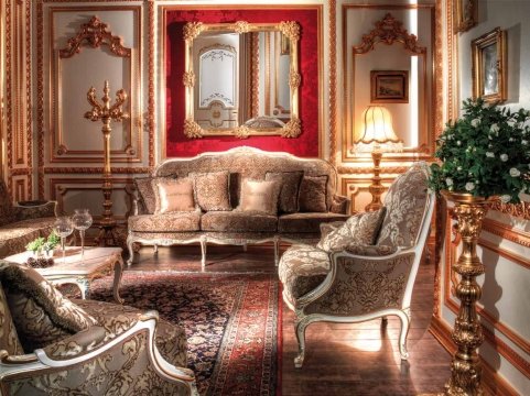 The picture shows a luxurious living room with a grand ceiling, crystal chandelier, and modern furnishings. The furniture pieces include a plush sofa, two armchairs, and an ottoman, all upholstered in ivory fabric. The walls are painted in a light shade of beige with ornate moldings around the ceiling and windows, creating a regal atmosphere. There is a unique chrome fireplace with a mantle adorned with several accessories, and a mirror is hung above it. A dark wooden grand piano stands against one wall, completing the stunning design.