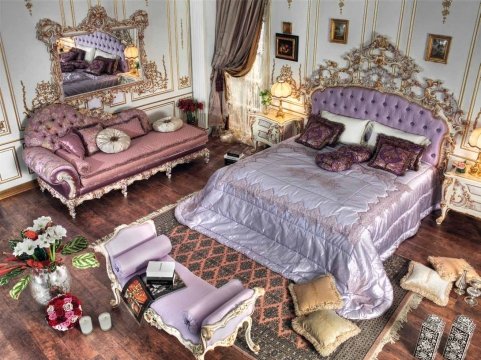 The picture shows an elegant living room with a modern, contemporary design. The room contains several pieces of furniture including two armchairs upholstered in a light grey fabric, a white sofa with decorative throw pillows, and a glass topped coffee table. The walls are painted a pale shade of blue and the floor is covered in a light colored rug. An ornate chandelier hangs from the ceiling, adding an upscale and luxurious touch to the room.