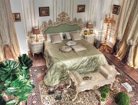This picture shows a beautiful and luxurious living room designed with a modern touch. The living room features a large cream-colored sofa with gold-colored accent pillows and two matching armchairs. There is a beige plush rug on the floor, and a large round black marble coffee table in front of the sofa. Behind it, there is an ornate silver fireplace surrounded by a wooden mantelpiece. The walls are painted in a soft beige color, and there is a large crystal chandelier hanging from the ceiling.