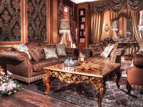 The picture shows an ornate hallway in a grand luxury home. It features a stunning interior design, with white marble floor and a prominent golden staircase with intricate handrails at the center. The high ceilings are adorned with luxurious chandeliers, and there are several mirrored surfaces that help to create a sense of space and grandeur. The walls are paneled in beige wood, with intricate sconces and floral artwork. The golden staircase leads up to a hallway with more exquisite detailing.