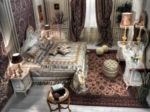 This picture shows an elegant and luxurious bedroom with a modern design. The room features light walls and dark wood flooring, and is decorated with golden accents. The centerpiece of the room is the large four-poster bed, with white and gold bedding draped over the frame. On either side of the bed are two nightstands, each with a lamp. A long bench is placed at the foot of the bed, and to the right is an armchair with a decorative ottoman. Large windows face out to the outside, allowing for plenty of natural light to come in.