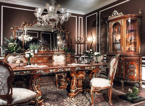 This picture shows a luxurious interior design. It features a lavish living space with a curved white sofa, brown wooden furniture and a white set of cabinets. The walls are beige with intricate gold accents and the floor is covered in an immaculate beige and white marble pattern. There is a decorative chandelier hanging from the ceiling, adding to the regal feel of the room.