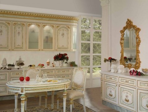 This picture shows a lavish Mediterranean style bedroom suite. It features a curved, button-tufted headboard with intricate carvings and details of cream, beige, and light brown tones. The bed is placed in front of two floor to ceiling windows with luxurious cream-colored drapes draped across them. On either side of the bed are two dark wood night stands with ornate lamps on top, and between them sits a large ottoman. The room is finished with a brown leather chaise lounge on one side and an elegant cream rug with brown detailing on the floor