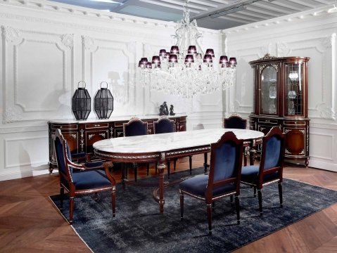 This picture shows an elegant, modern-style dining room with two red velvet armchairs situated opposite a light-colored glass table. There is a large silver-colored chandelier hanging from the ceiling and several light-colored curtains along the walls. The floor of the room is covered in tile, and a luxurious rug rests in the center of the space.